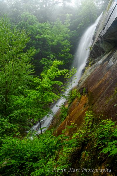 Cascade Falls in Vermont with green foliage and red rock in the foreground, and fog in the background.