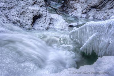 Framed by sparkling ice, the lower tier of Moss Glen Falls in Stowe Vermont flows down towards a branch covered in icicles and out through the snowy ravine walls. The color of the refreezing water is a bluish green.